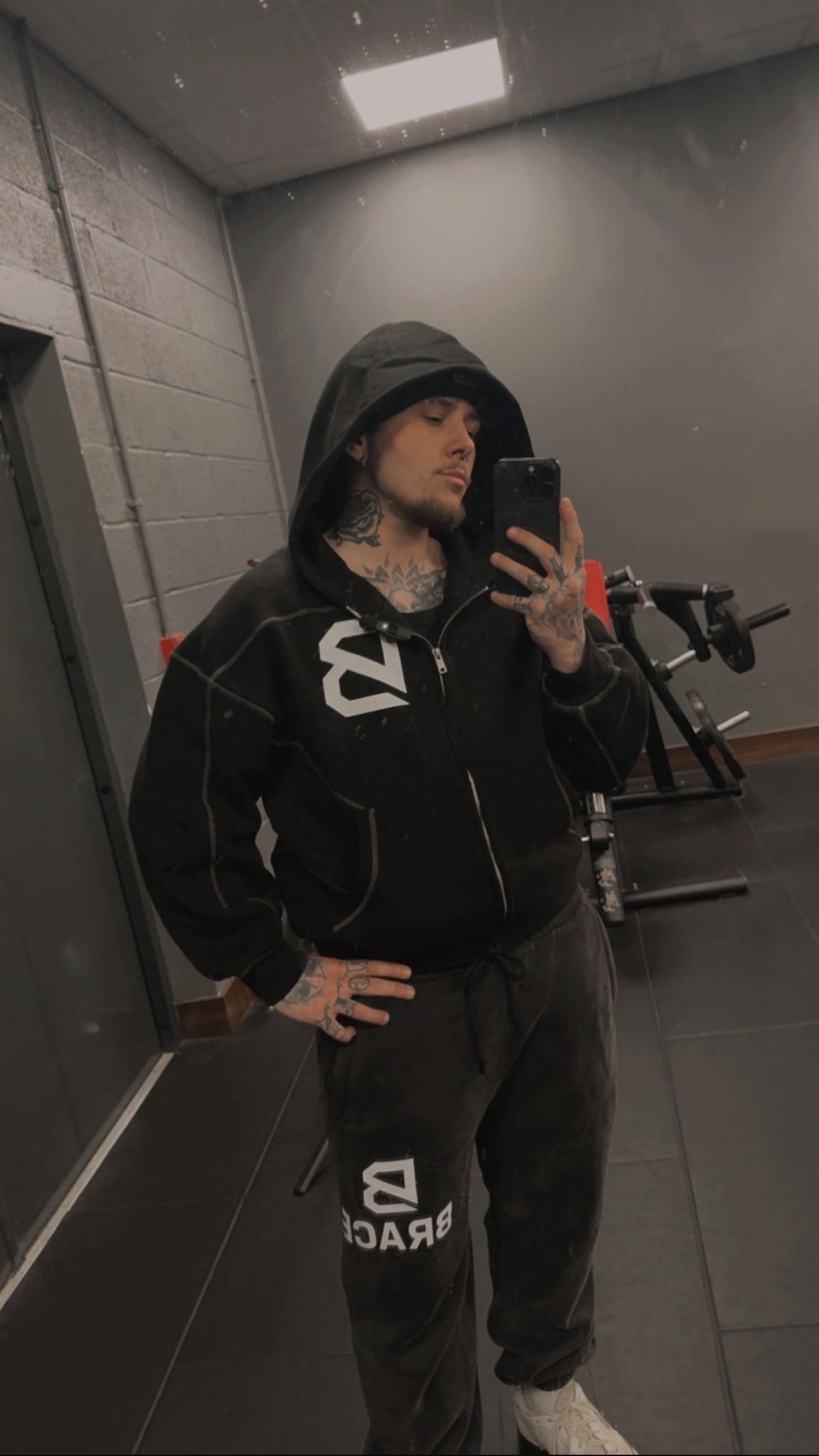 Harry wearing BRACE hoodie and joggers in the gym, showcasing the brand's bold logo on his chest and leg. The photo captures him standing confidently in a gym mirror.