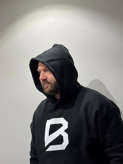 Ade Mills, owner of BRACE GymWear, wearing a black BRACE hoodie with the white logo, looking thoughtful against a plain wall, symbolizing resilience and mental strength.